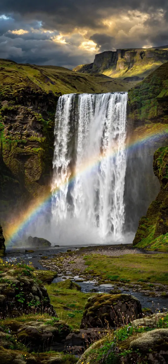 Tall waterfall with a rainbow, green cliffs and mossy rocks under cloudy sky